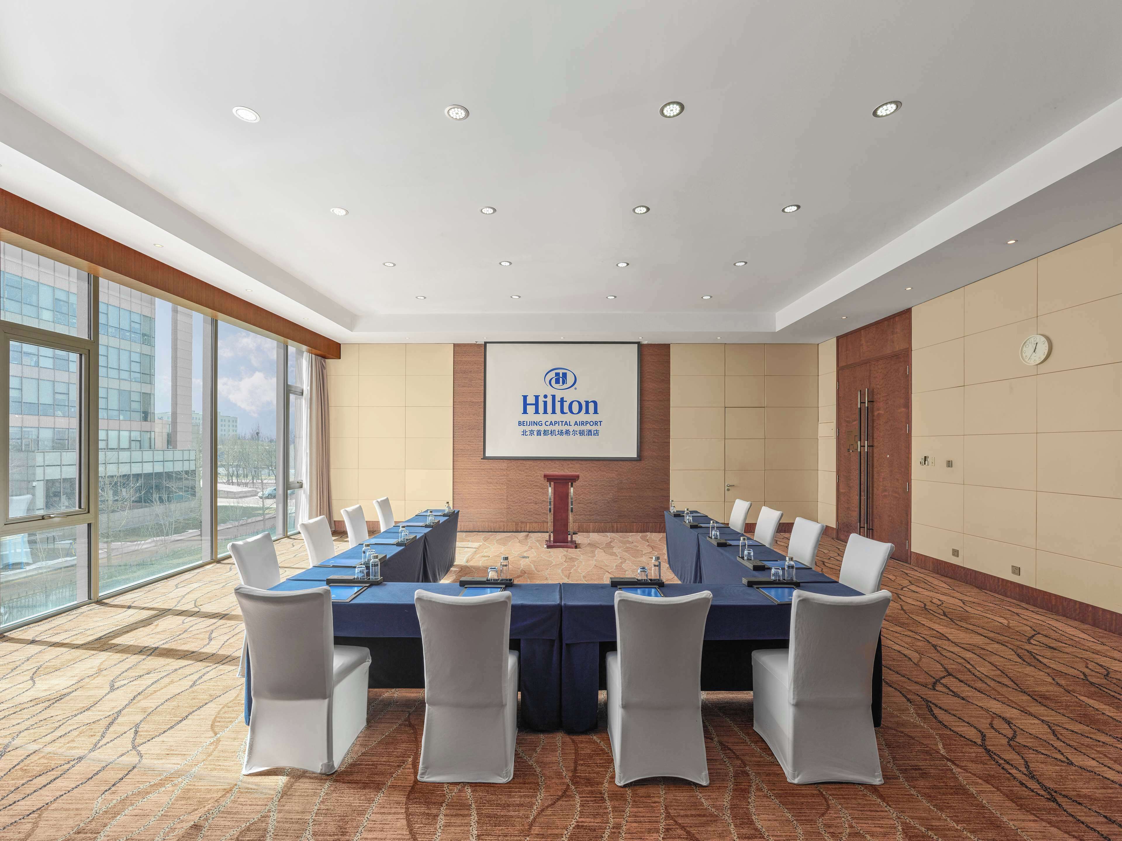 A conference room with a long table, white chairs, a podium, and a screen displaying the Hilton logo.
