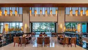 A spacious hotel lobby with multiple dining tables and chairs, large windows offering a view of the outdoors, and a modern ceiling design with hanging lights.