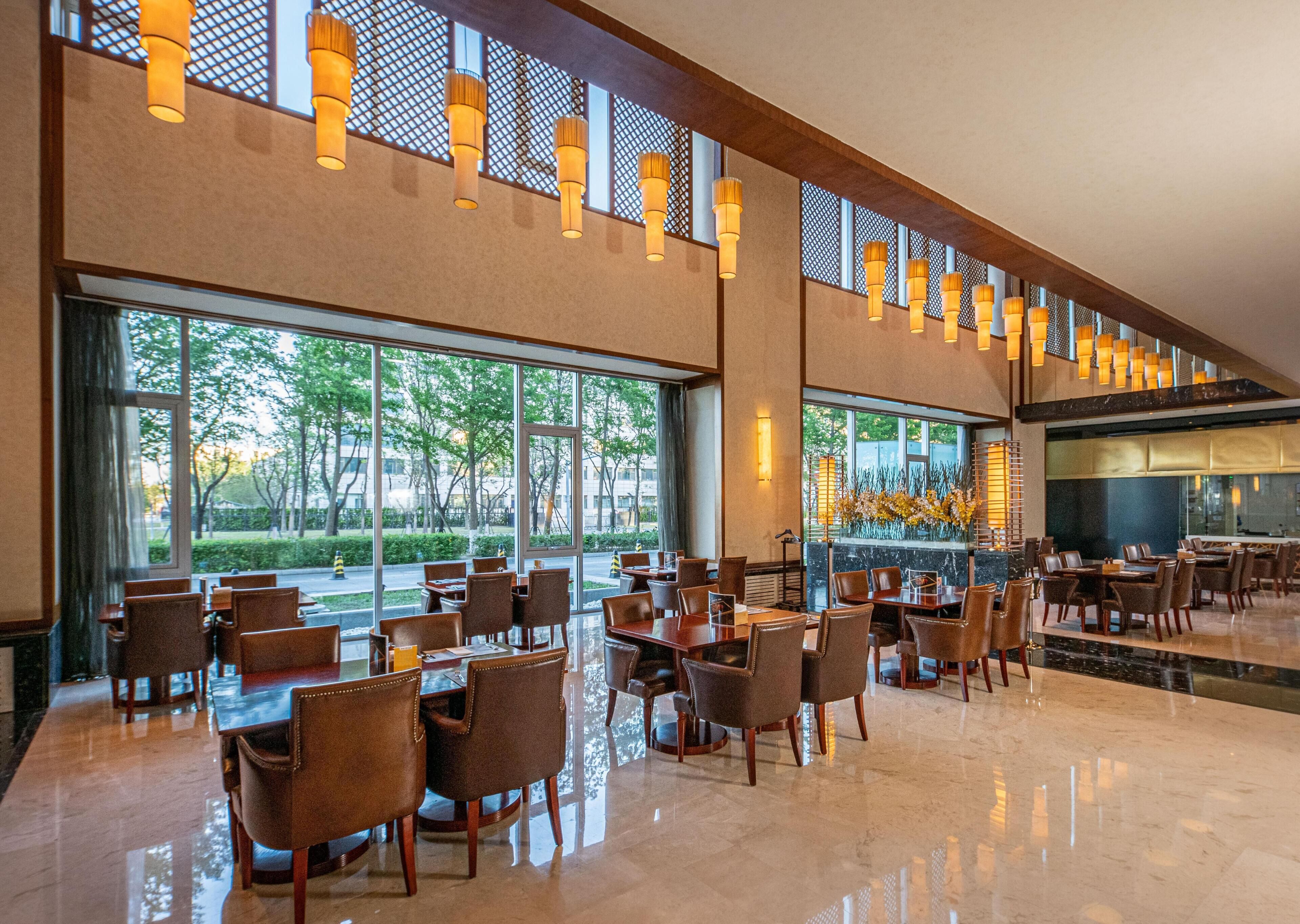 A spacious indoor area with multiple tables and chairs, large windows offering a view of the outdoors, and a modern ceiling design with hanging lights.