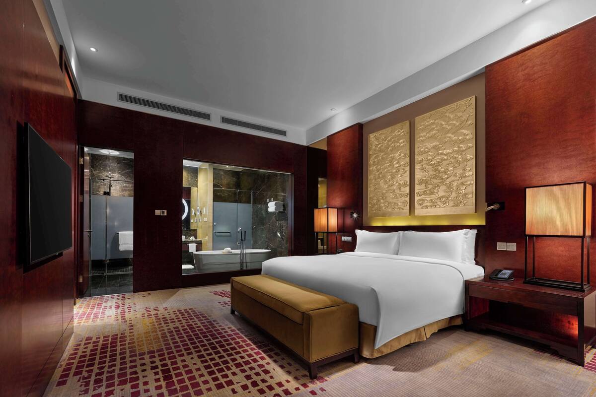 Una habitación de hotel moderna con una cama grande, una mesita de noche con una lámpara, una mesita más pequeña con un teléfono, un espejo grande y una puerta corredera que da a un baño.
