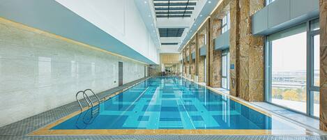 An indoor swimming pool with clear blue water, a tiled floor, and large windows on one side.