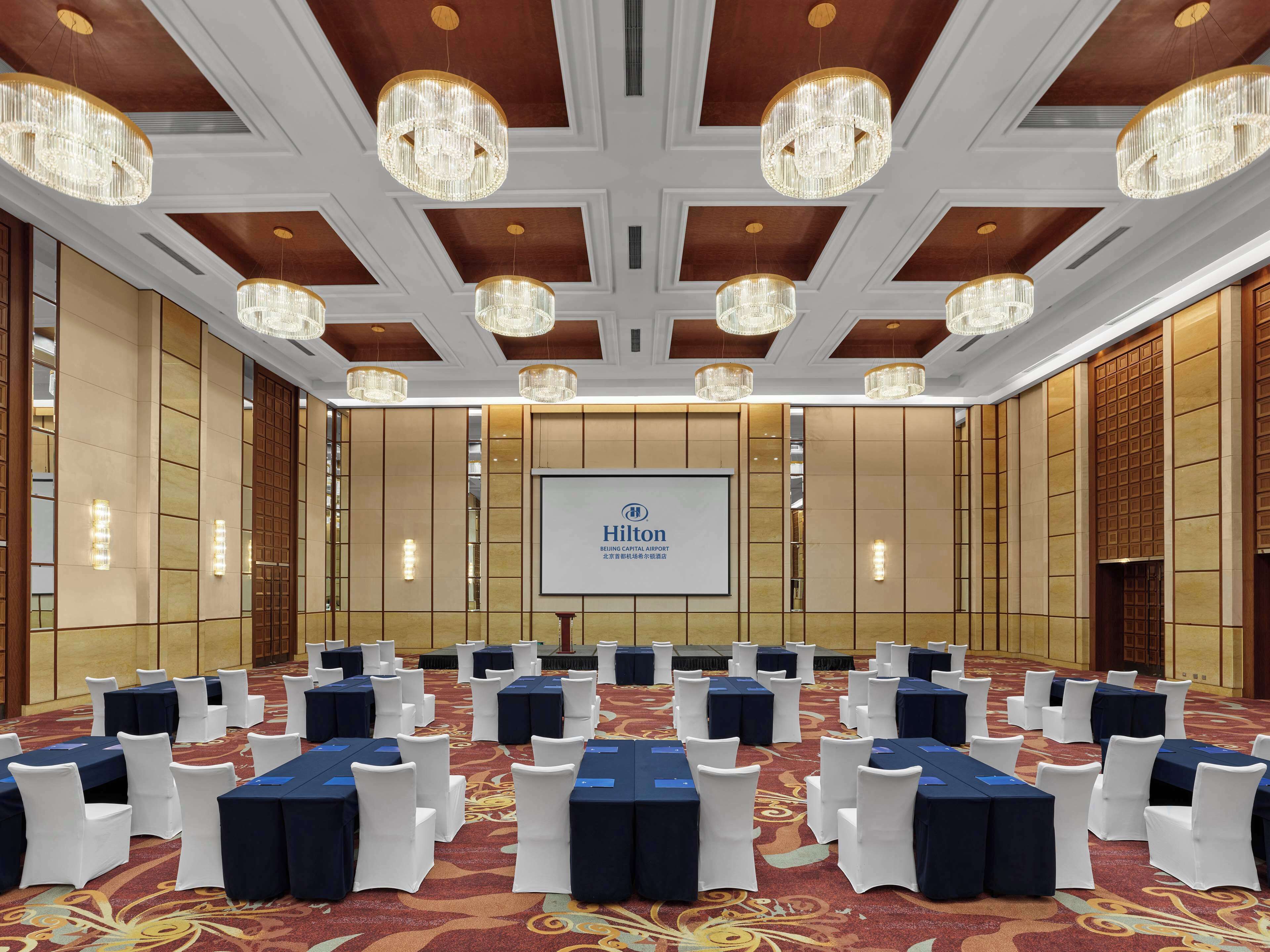 A conference room with rows of tables and chairs, chandeliers, and a screen displaying the Hilton logo.