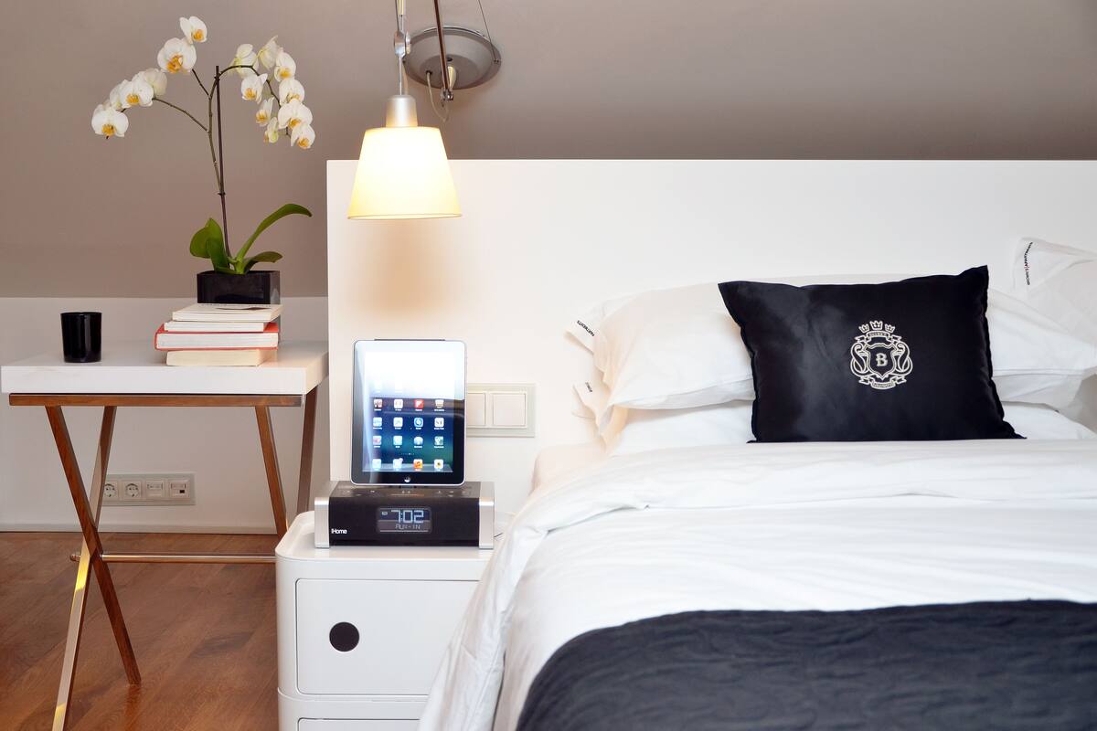 Une chambre d’hôtel bien rangée, avec un lit, une table de chevet, une lampe et un petit bureau agrémenté d’une plante et de quelques livres.