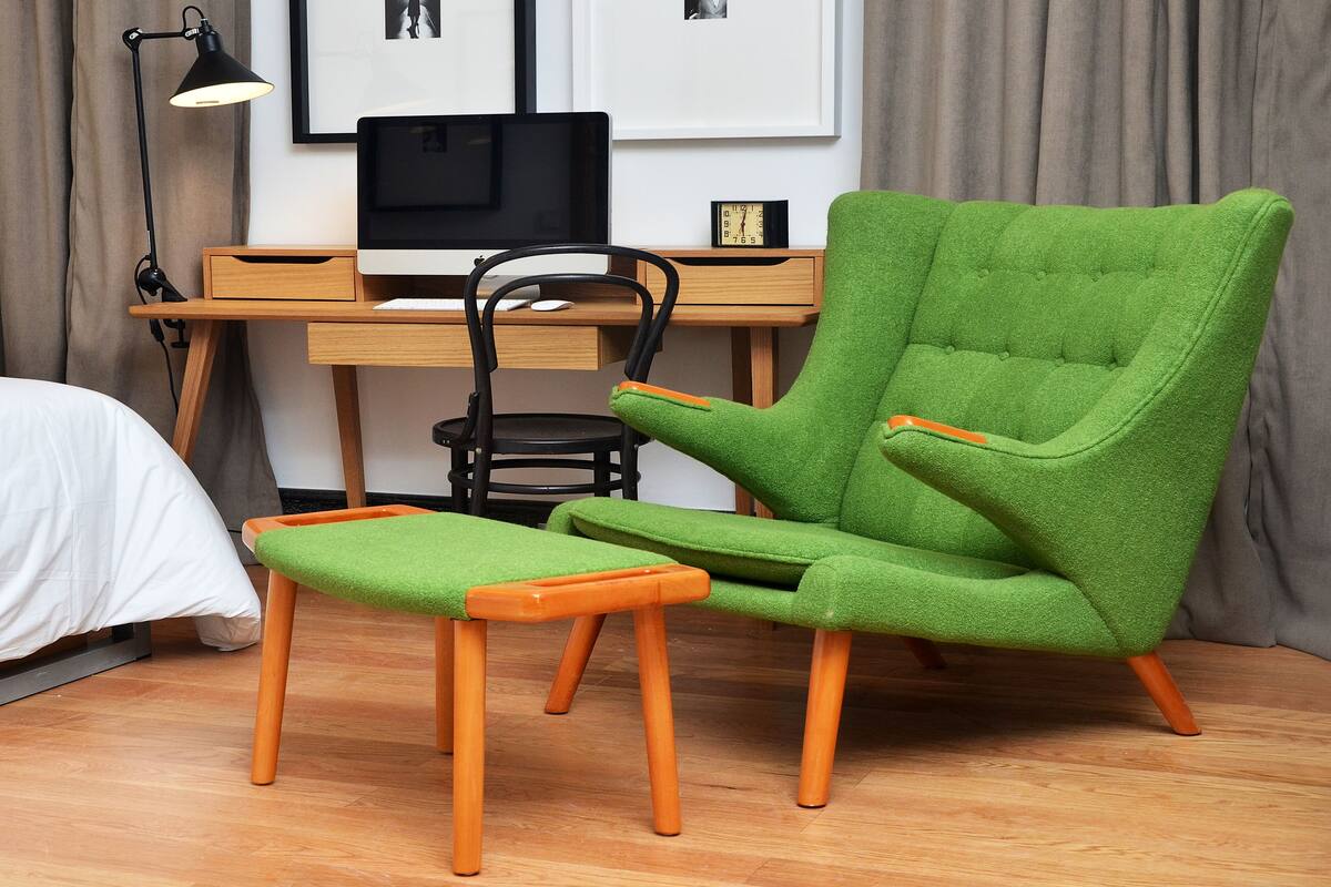 Une pièce comprenant un fauteuil vert, un pouf assorti, un bureau en bois, une chaise noire, une lampe de bureau noire et un tableau encadré accroché au mur.