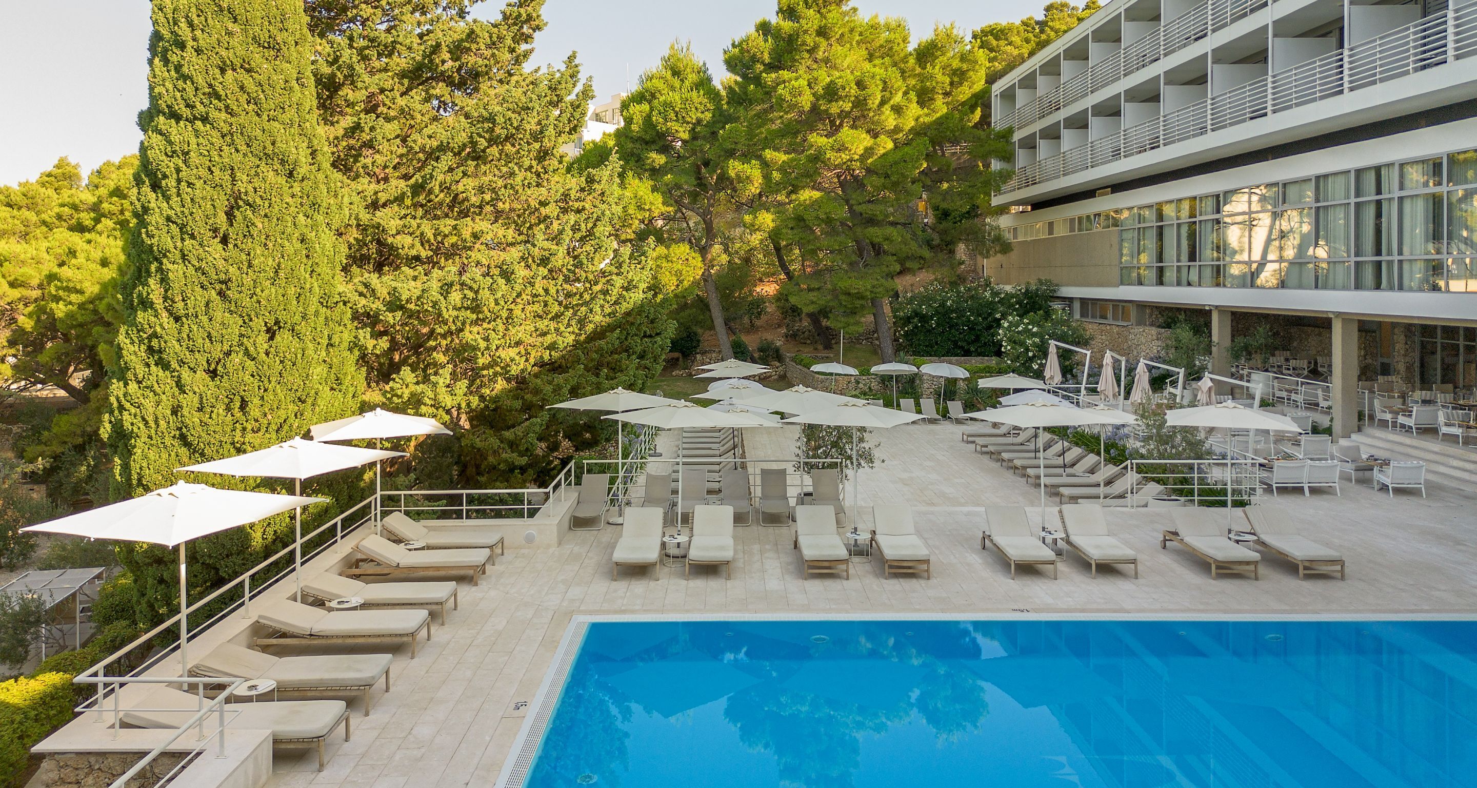 A pool area with sun loungers and umbrellas, surrounded by trees and a modern building.