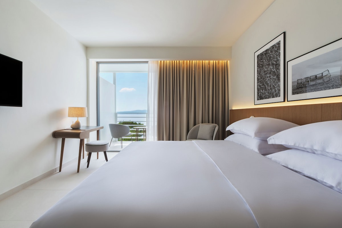 A hotel room with a large bed, a desk with a lamp, a chair, and a view of the outdoors.