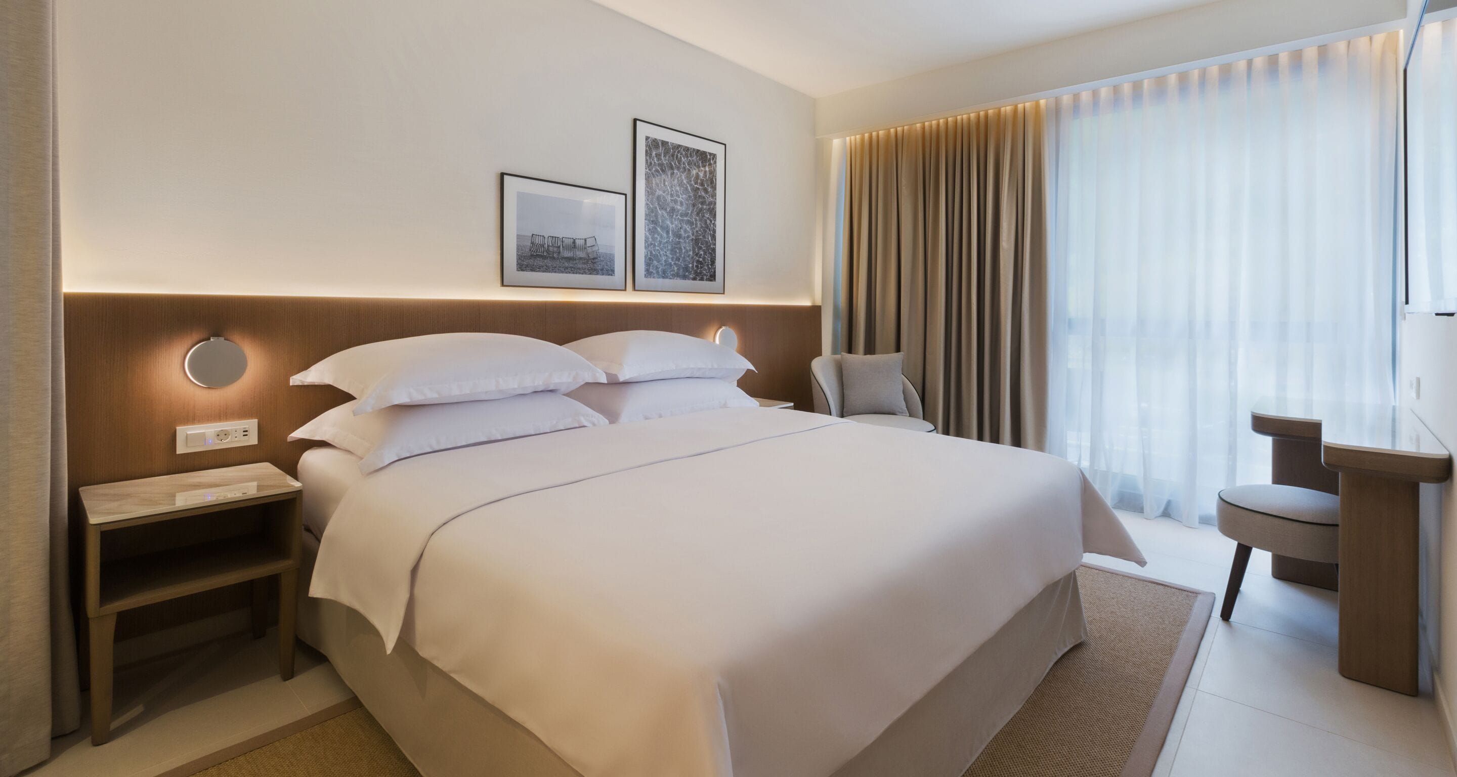 Quarto de hotel moderno com cama grande, mesas de cabeceira, escrivaninha e vista para o exterior através de cortinas translúcidas.