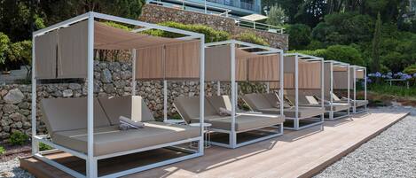 Fila de sillones de exterior con toldos, sobre una terraza de madera con un muro de piedra y vegetación al fondo.