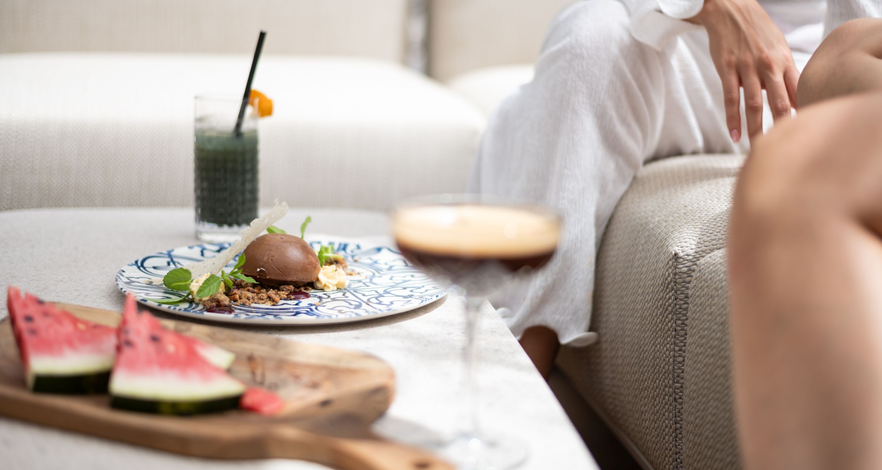 A dining area with a plate of food, a glass of drink, and a wooden board with watermelon slices.