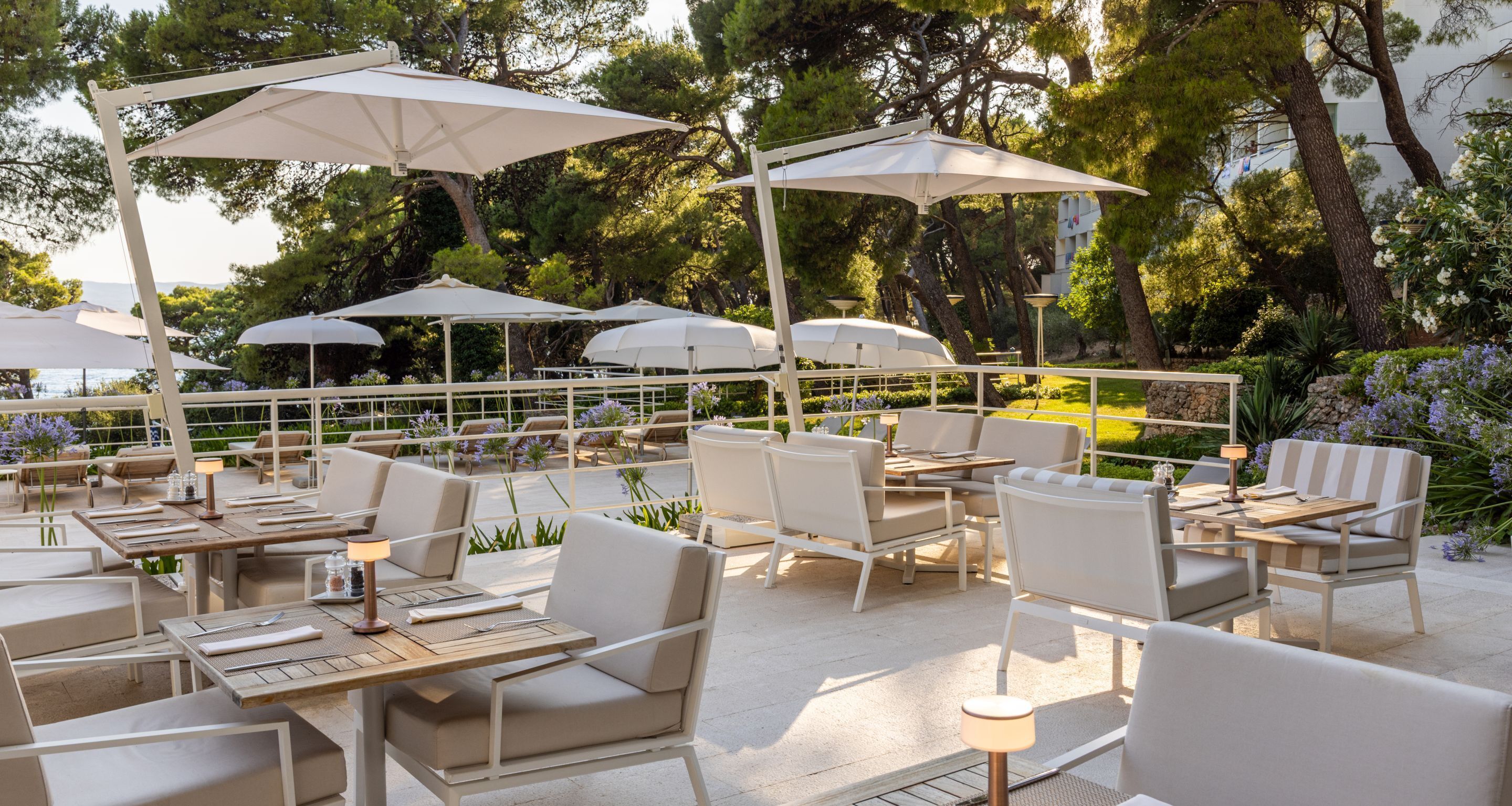An outdoor patio with white furniture, umbrellas, and a view of trees and a building in the background.