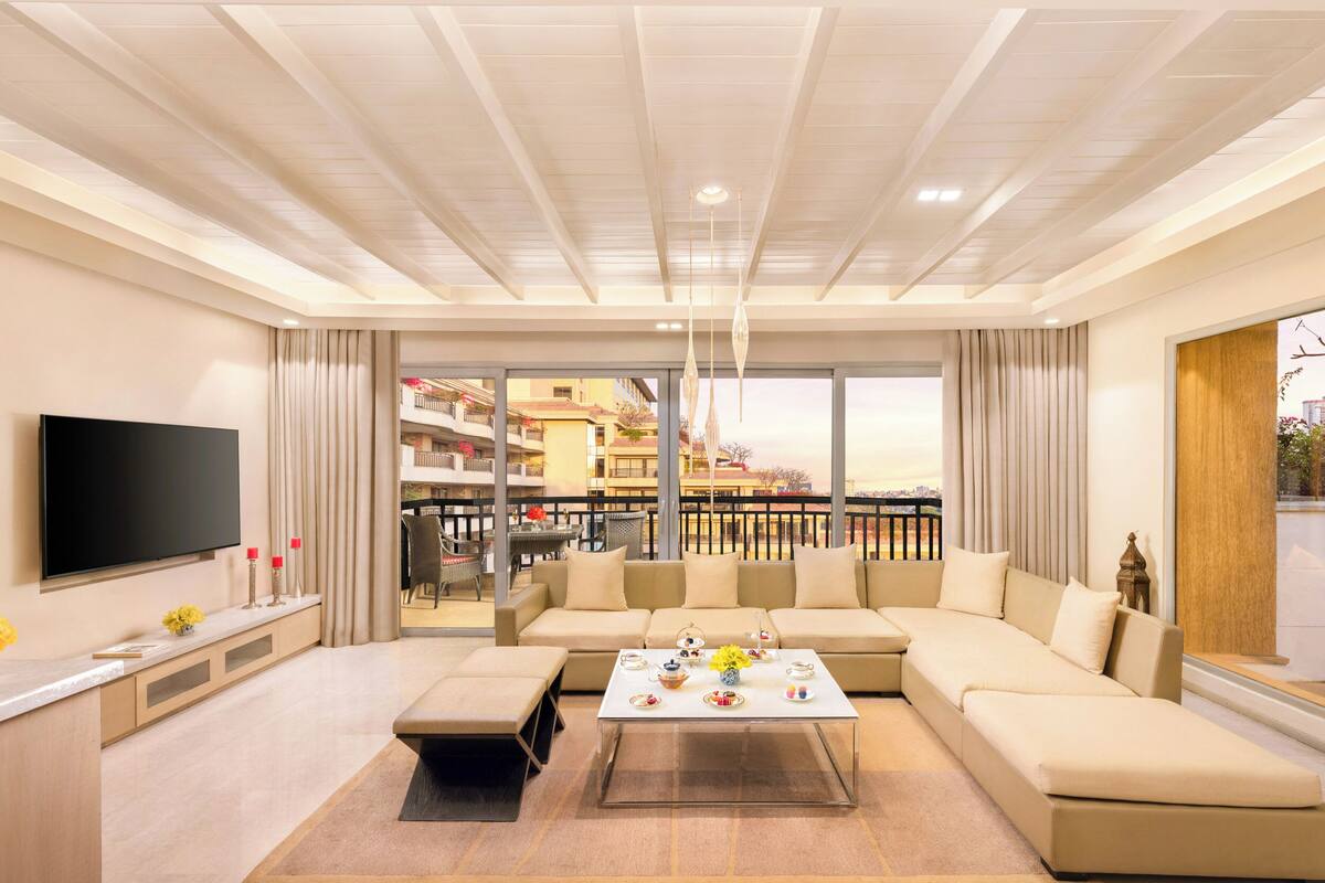 A modern living room with a large sectional sofa, a flat-screen TV, a coffee table with decorative items, and a balcony with a view of the city.