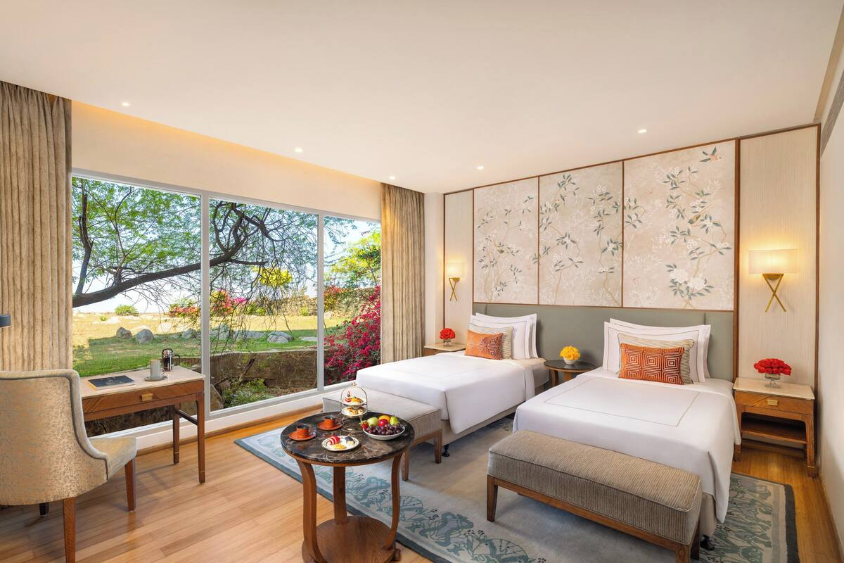 A hotel room with two beds, a desk, a table with food, and large windows with a view of greenery.