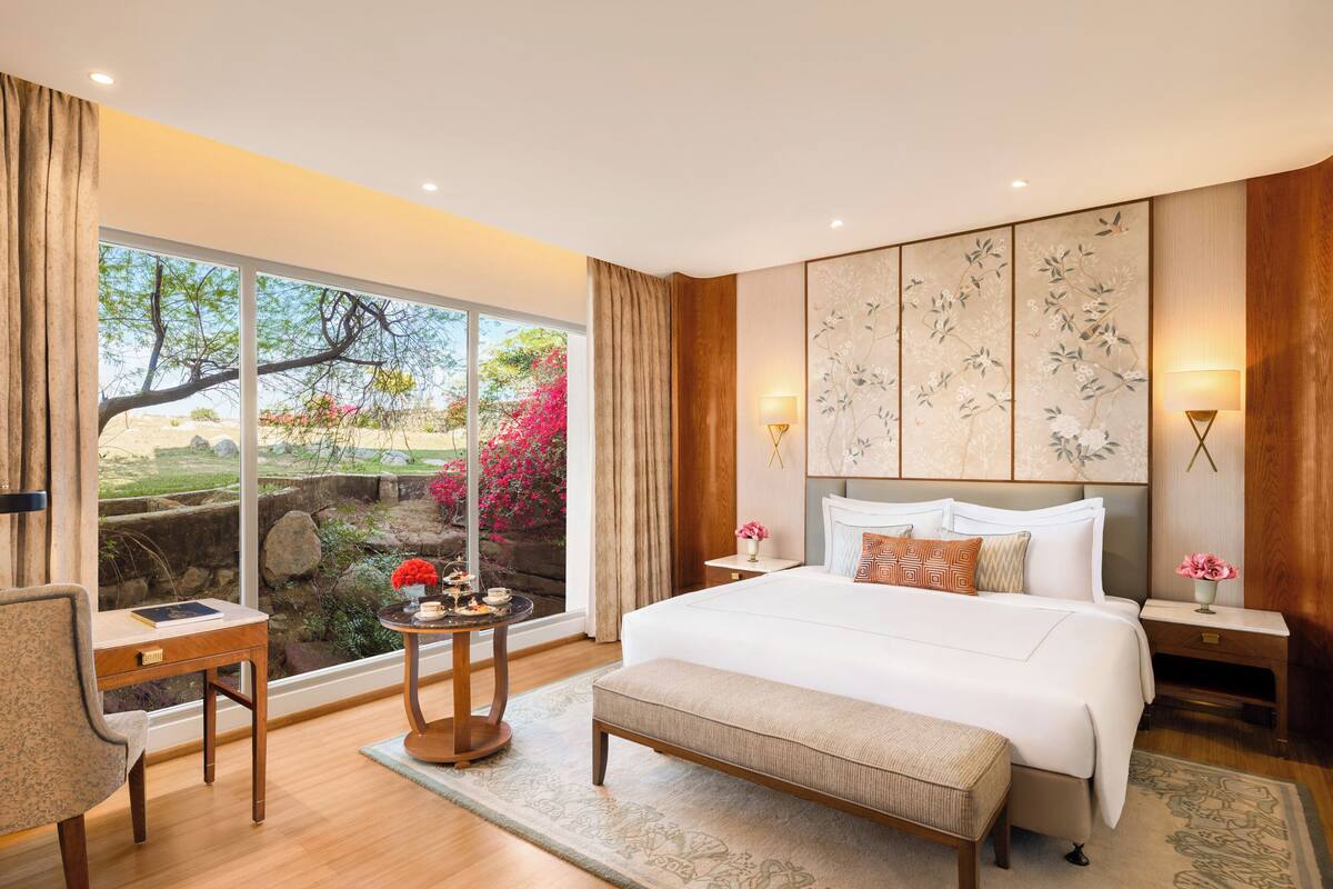 A spacious bedroom with a large bed, a desk, and a view of a garden through floor-to-ceiling windows.