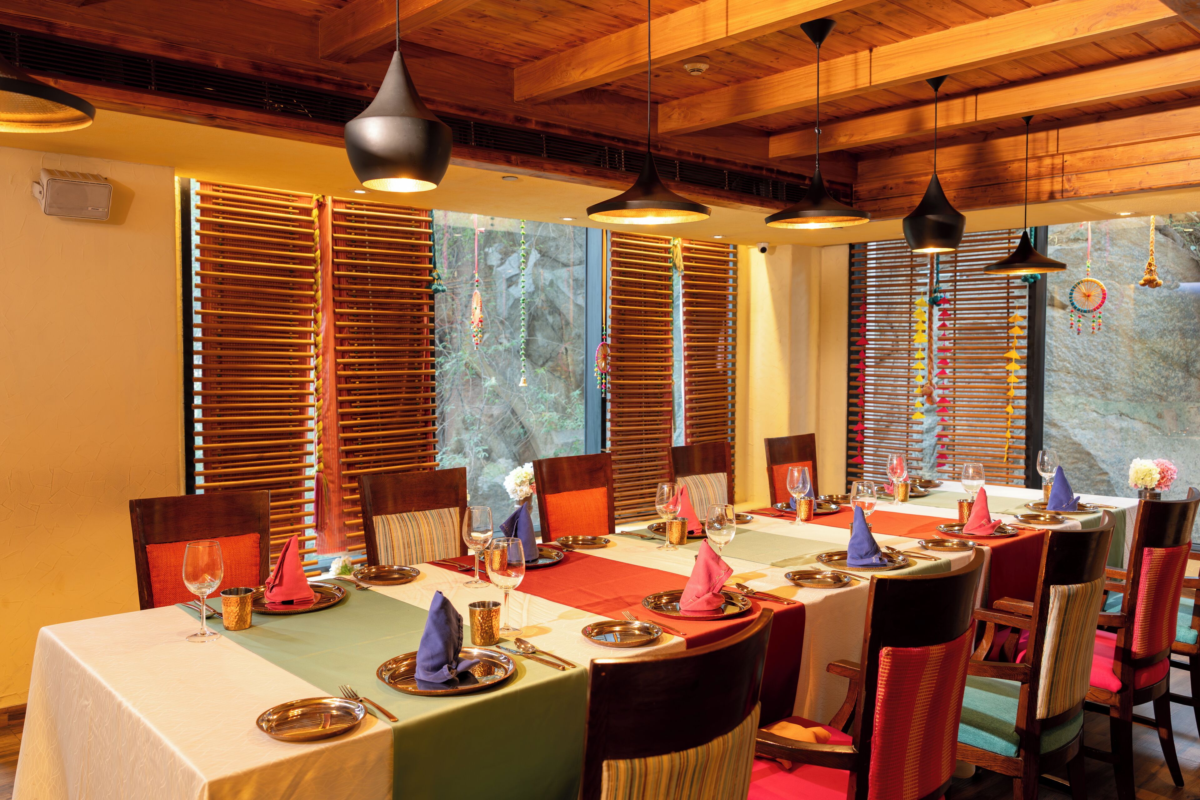 A dining area with a long table set for a meal, wooden ceiling, and large windows with blinds.