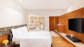 A modern hotel room with a large bed, a flat-screen TV, wooden paneling, and a bathroom visible through a glass door.