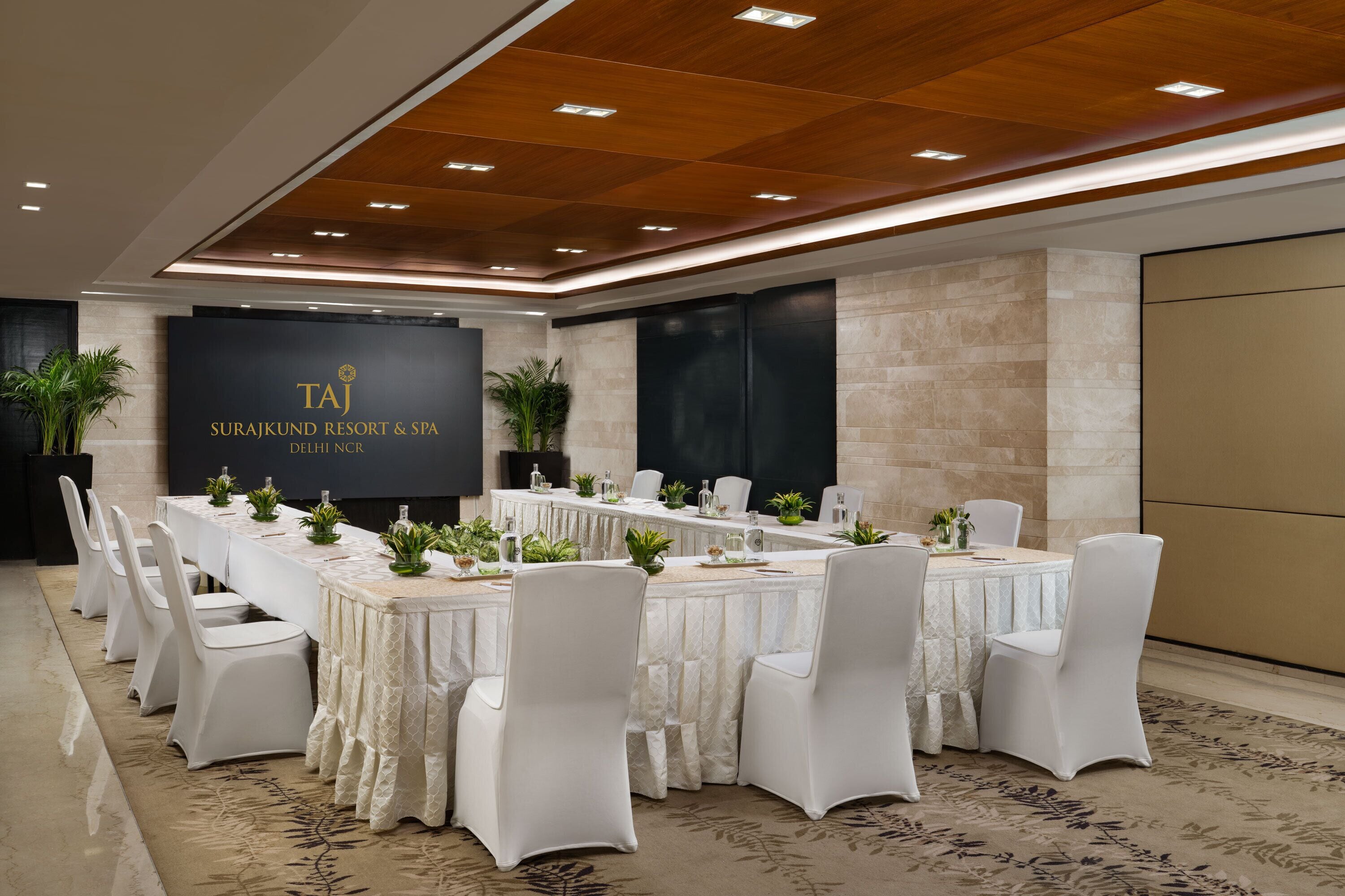 Een vergaderruimte met een lange tafel, witte stoelen en een achtergrond met het logo van Taj Suryakund Resort & Spa.