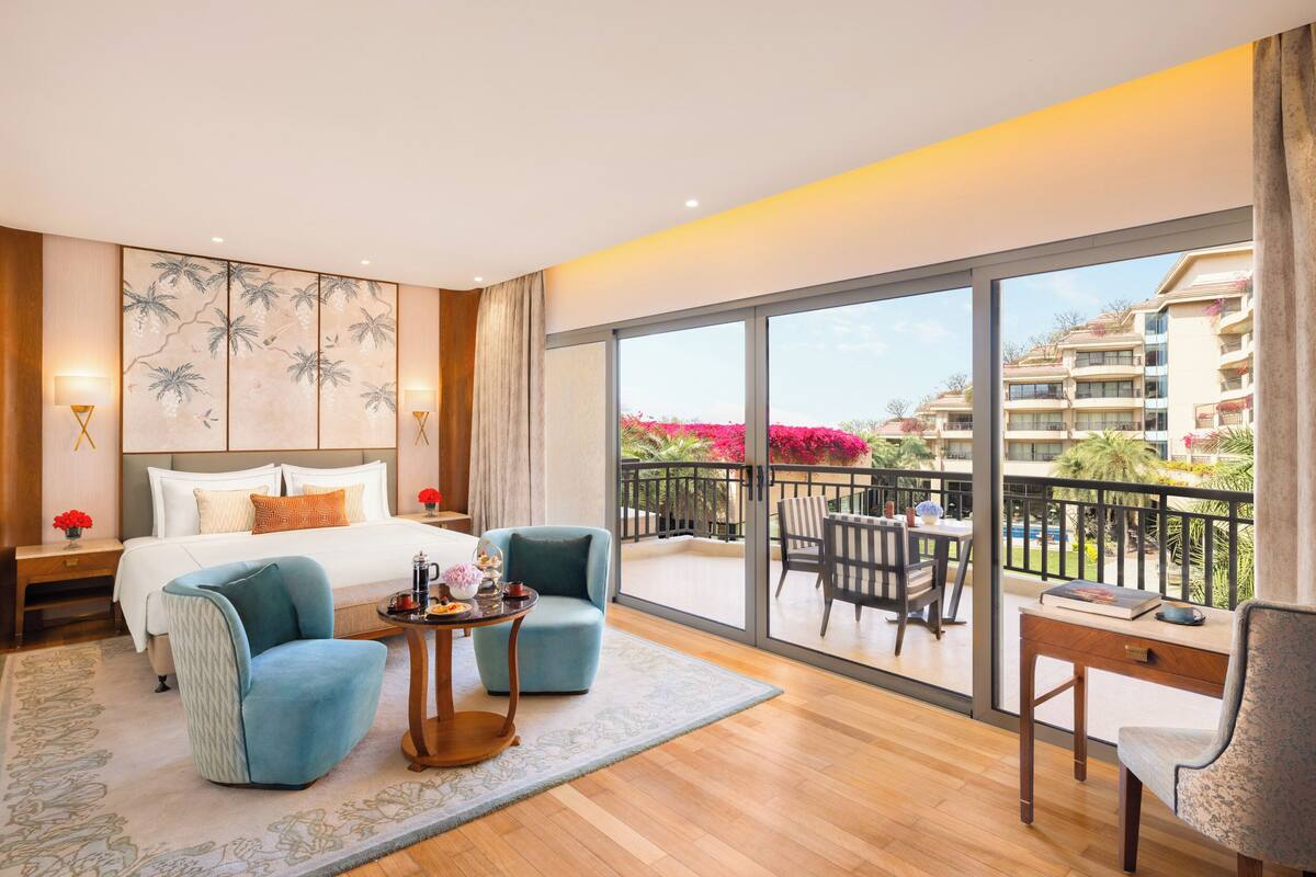 A hotel room with a large bed, two armchairs, a small table, and a balcony with a view of the hotel and surrounding area.