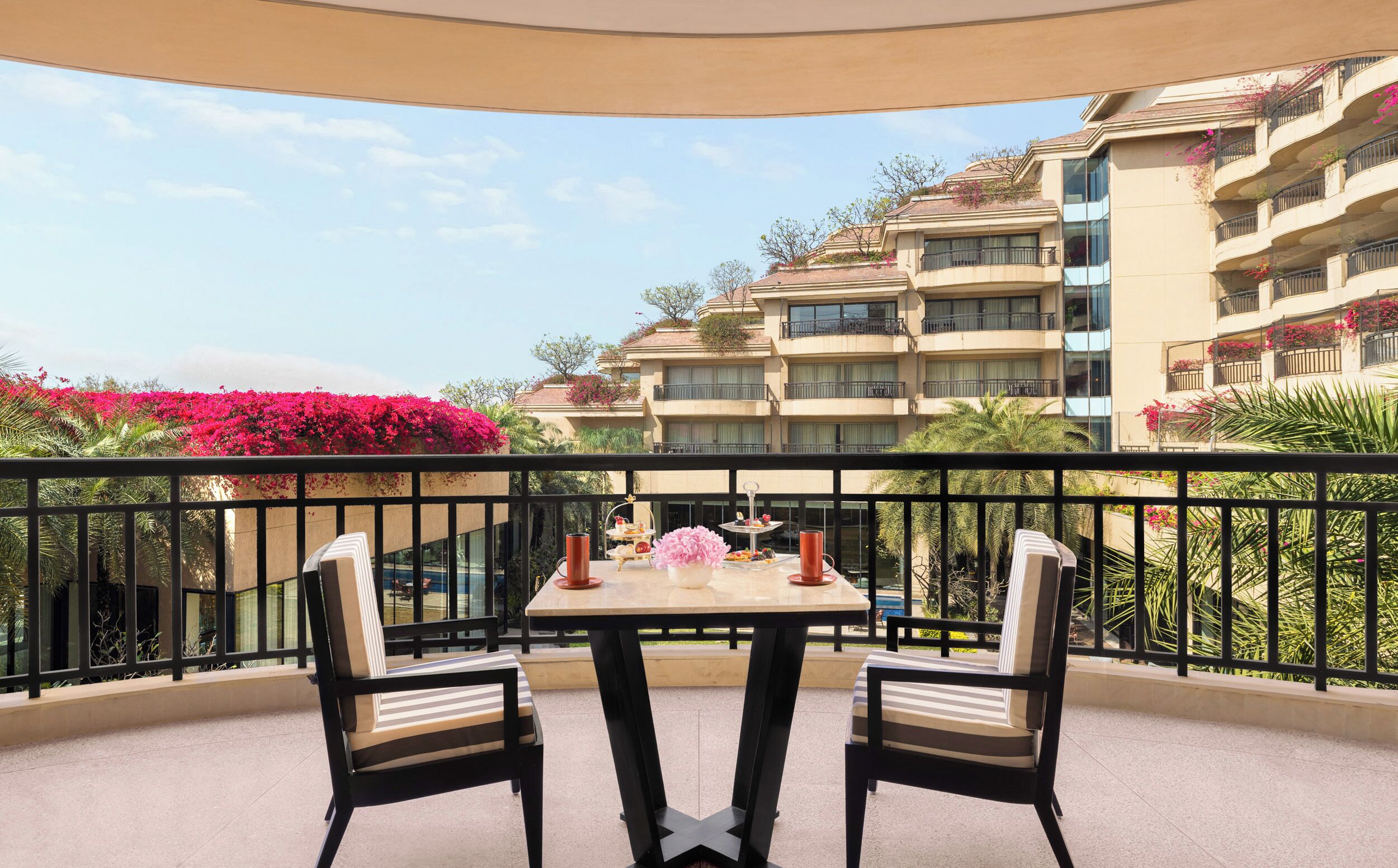 A balcony with two chairs and a table, overlooking a building with balconies and red flowers.