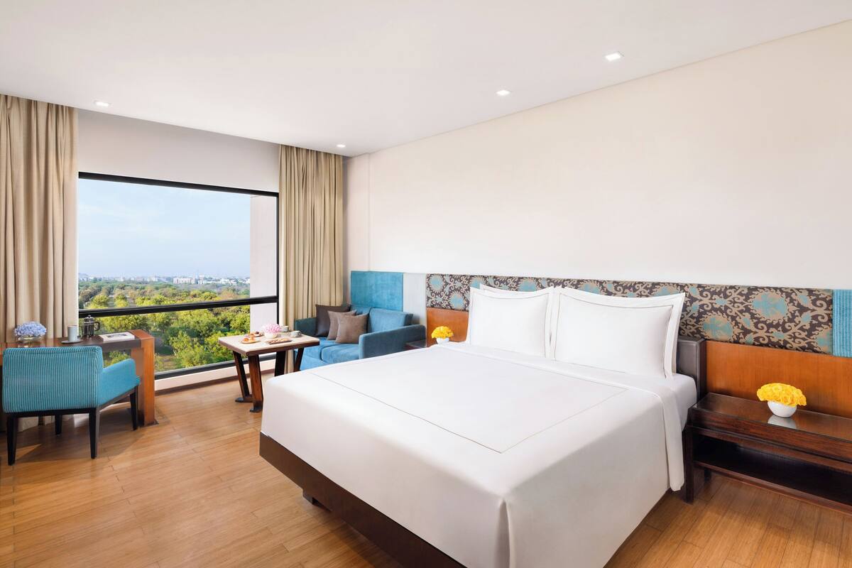 A hotel room with a large bed, a desk, a chair, and a view of the city through a large window.