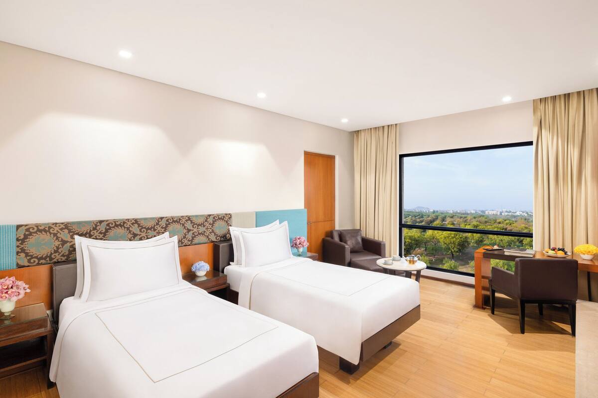 A hotel room with two beds, a desk, a chair, and a large window offering a city view.