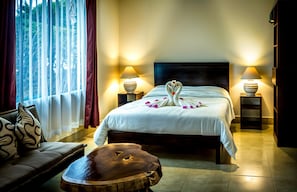 A hotel room with a large bed, two bedside lamps, a wooden coffee table, a sofa, and a view of trees through the window. - La Villa Creole (Jacó)