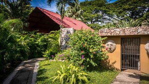 A tropical garden with a stone wall, a red-roofed structure, and a small building with a barred door. - La Villa Creole (Jacó)