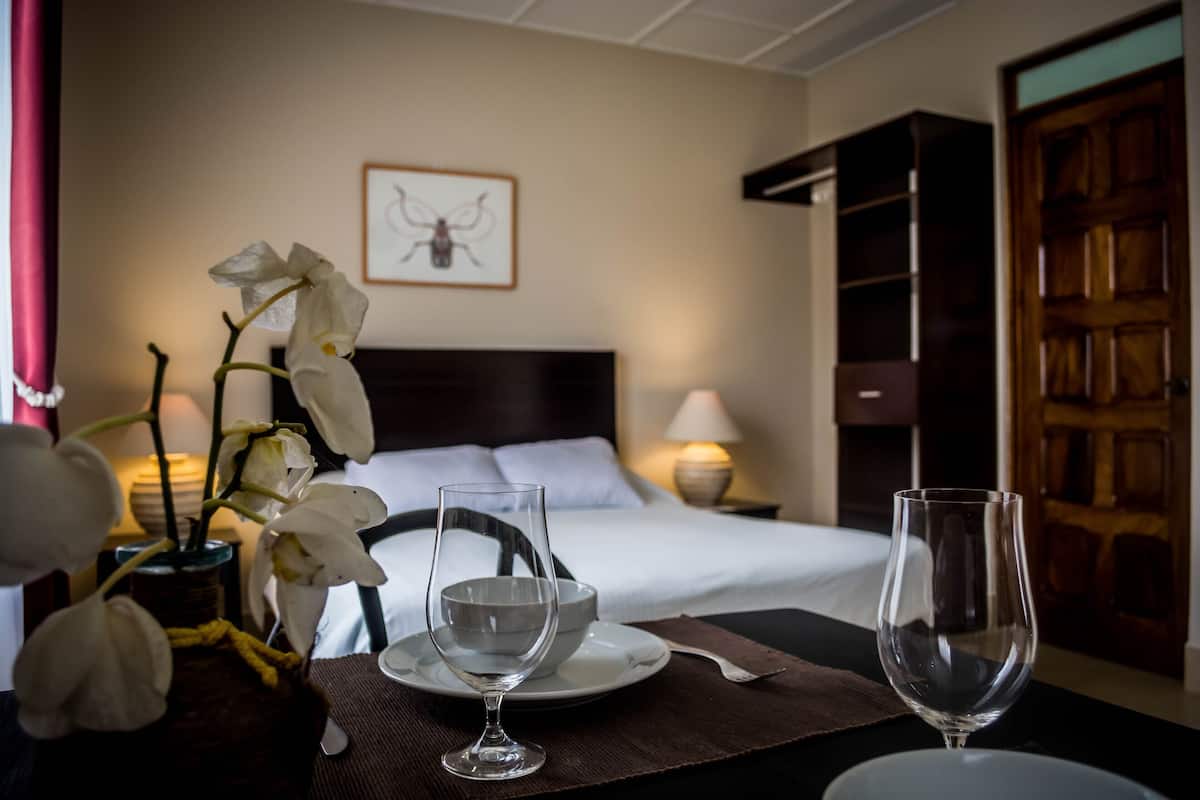 A hotel room with a bed, a bedside table with a vase of white flowers, two empty wine glasses, and a framed picture on the wall.