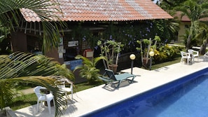 A poolside area with lounge chairs, a reception desk, and a building with a tiled roof. - La Villa Creole (Jacó)