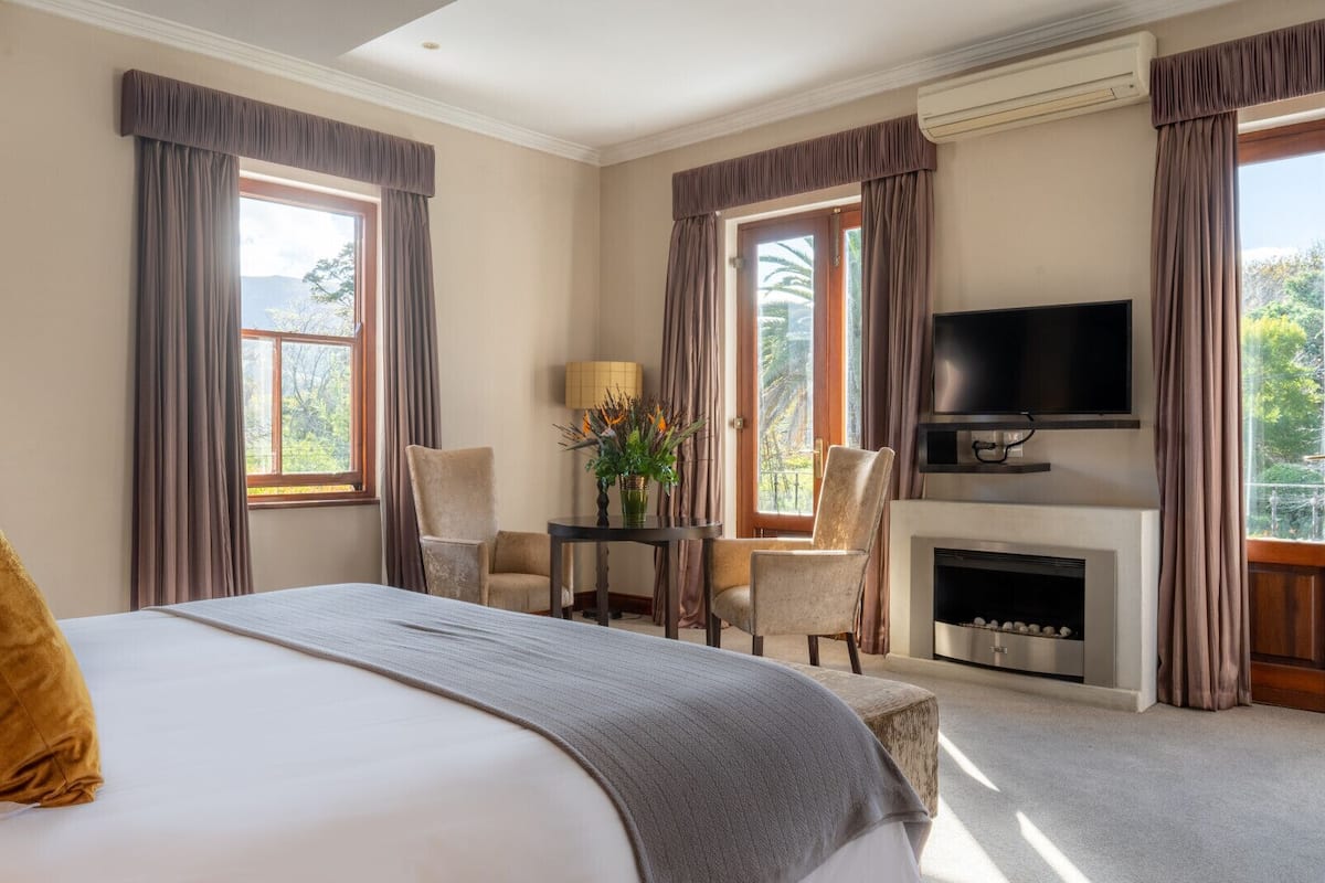 A hotel room with a large bed, a television, a fireplace, and a view of trees through the windows.