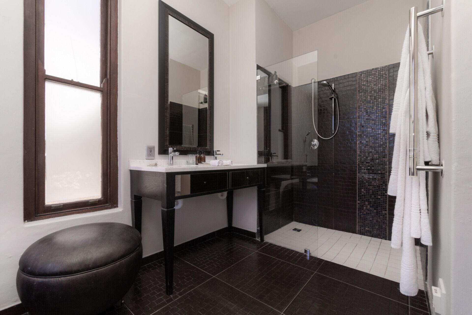 Une salle de bain moderne avec une douche carrelée aux couleurs sombres, un grand miroir et un tabouret en cuir noir.