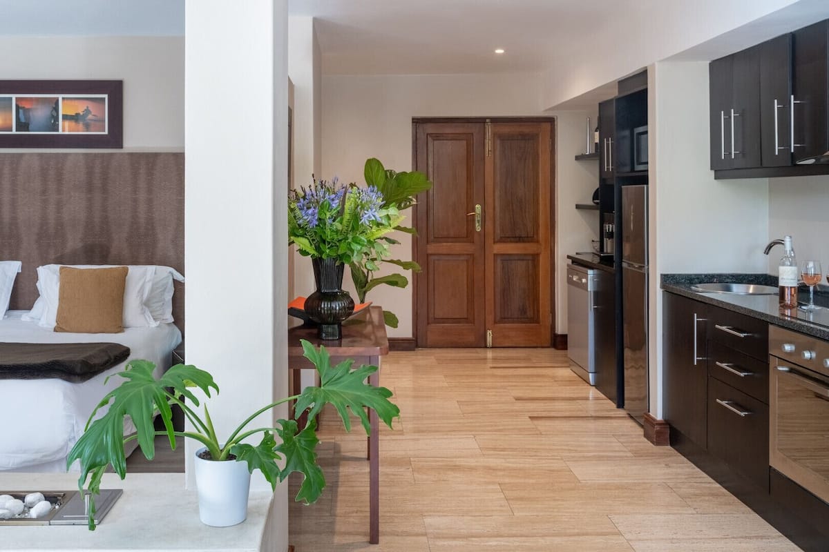 A modern hotel room with a bed, a small table with plants, a kitchen area with dark cabinets and a stainless steel refrigerator.
