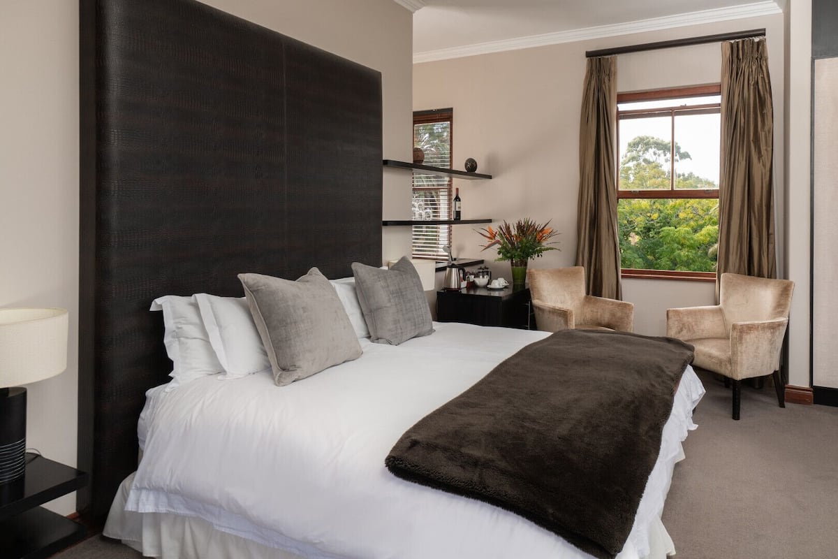 A hotel room with a large bed, a bedside table with a lamp, a chair, a window with curtains, and a shelf with decorative items.