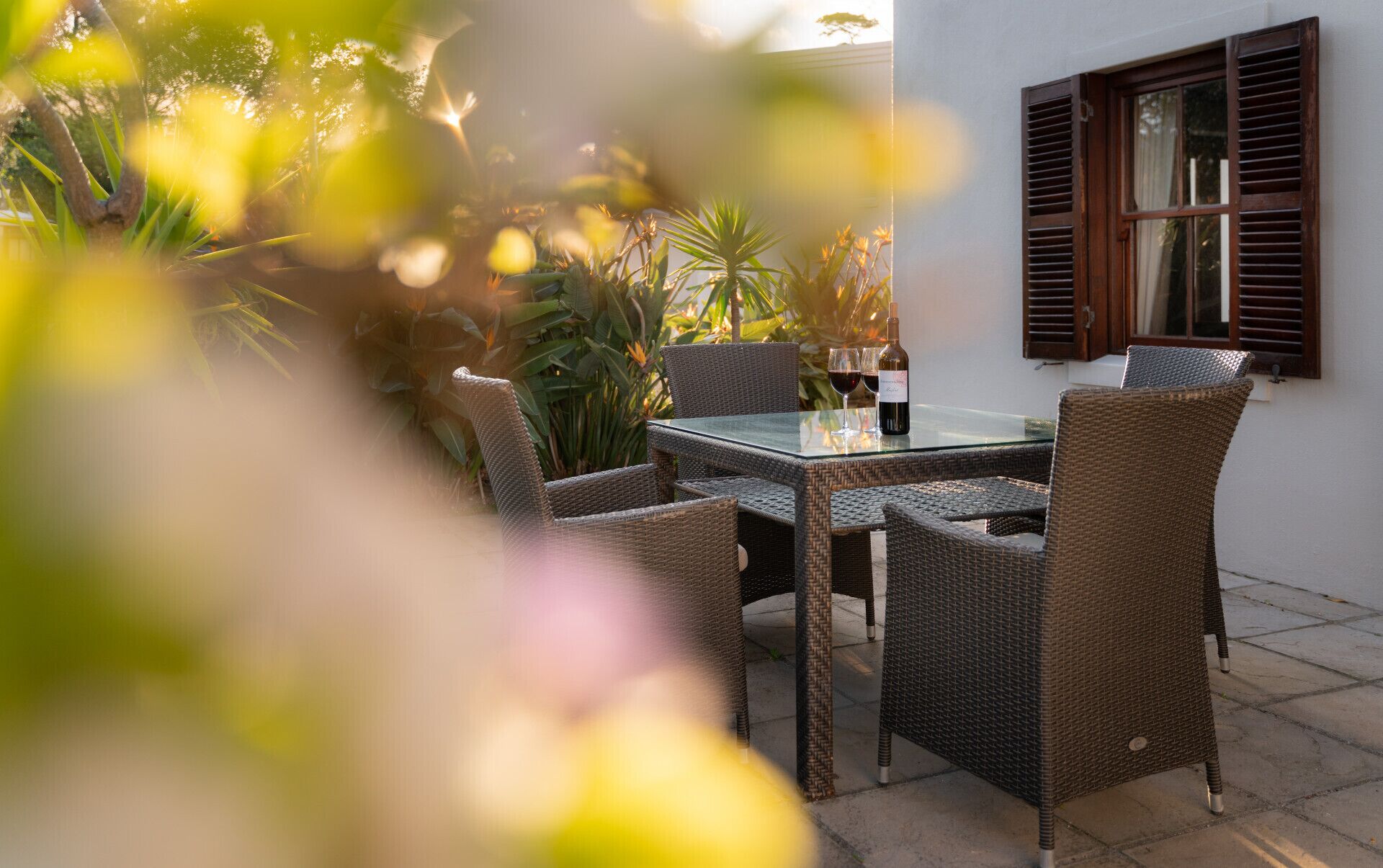 An outdoor dining area with wicker chairs and a glass table, surrounded by plants and a window with shutters.