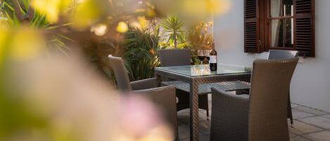 An outdoor dining area with wicker chairs and a glass table, surrounded by plants and a window with shutters.