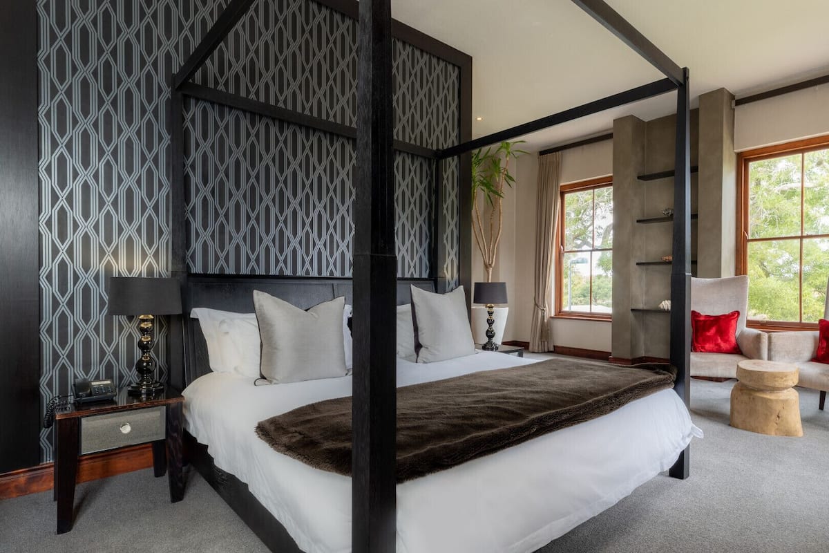 A four-poster bed with a dark headboard, a bedside table with a lamp, a chair with a red cushion, and a floor lamp.