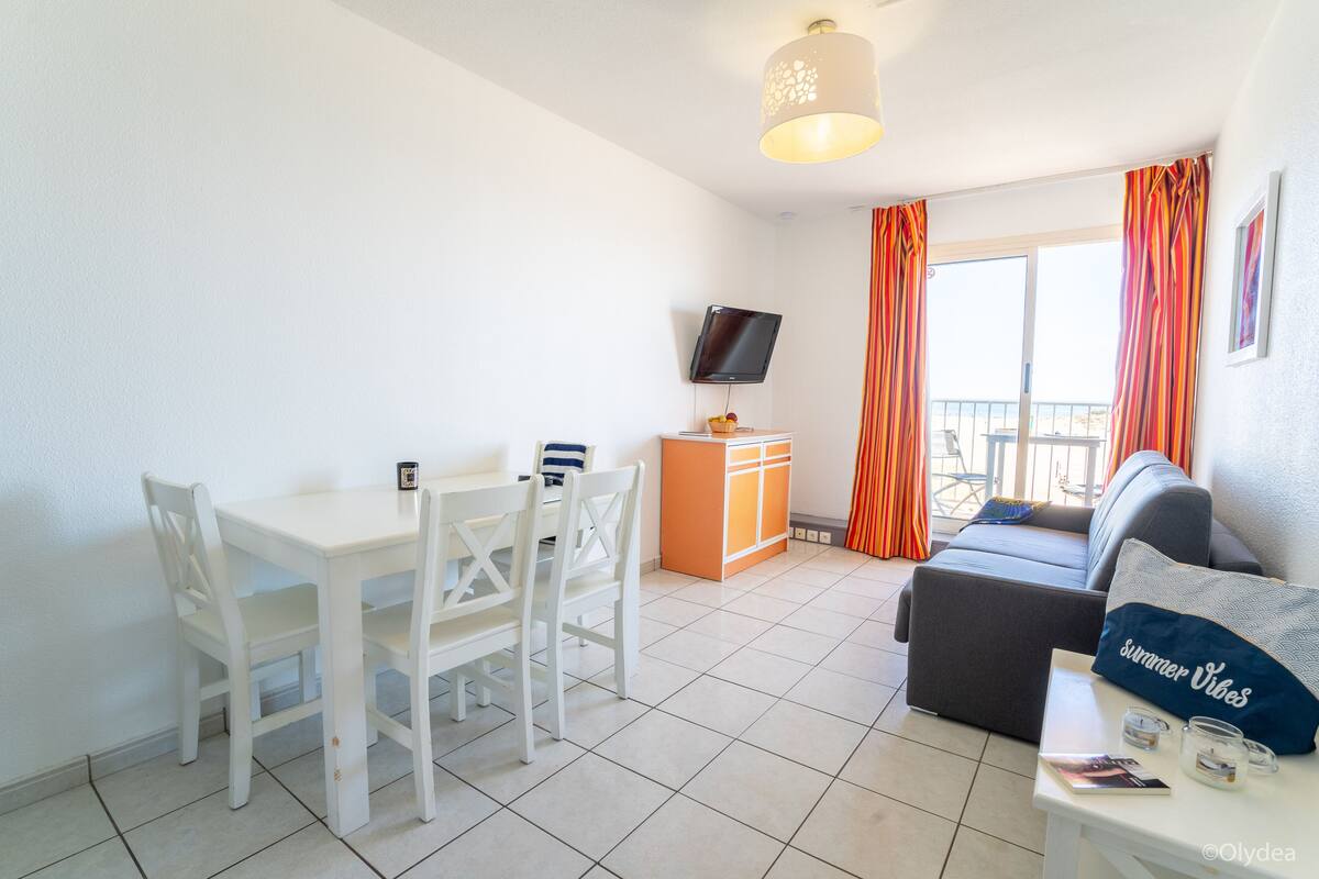 A room with a dining table, chairs, a sofa, a TV, and a balcony with curtains.