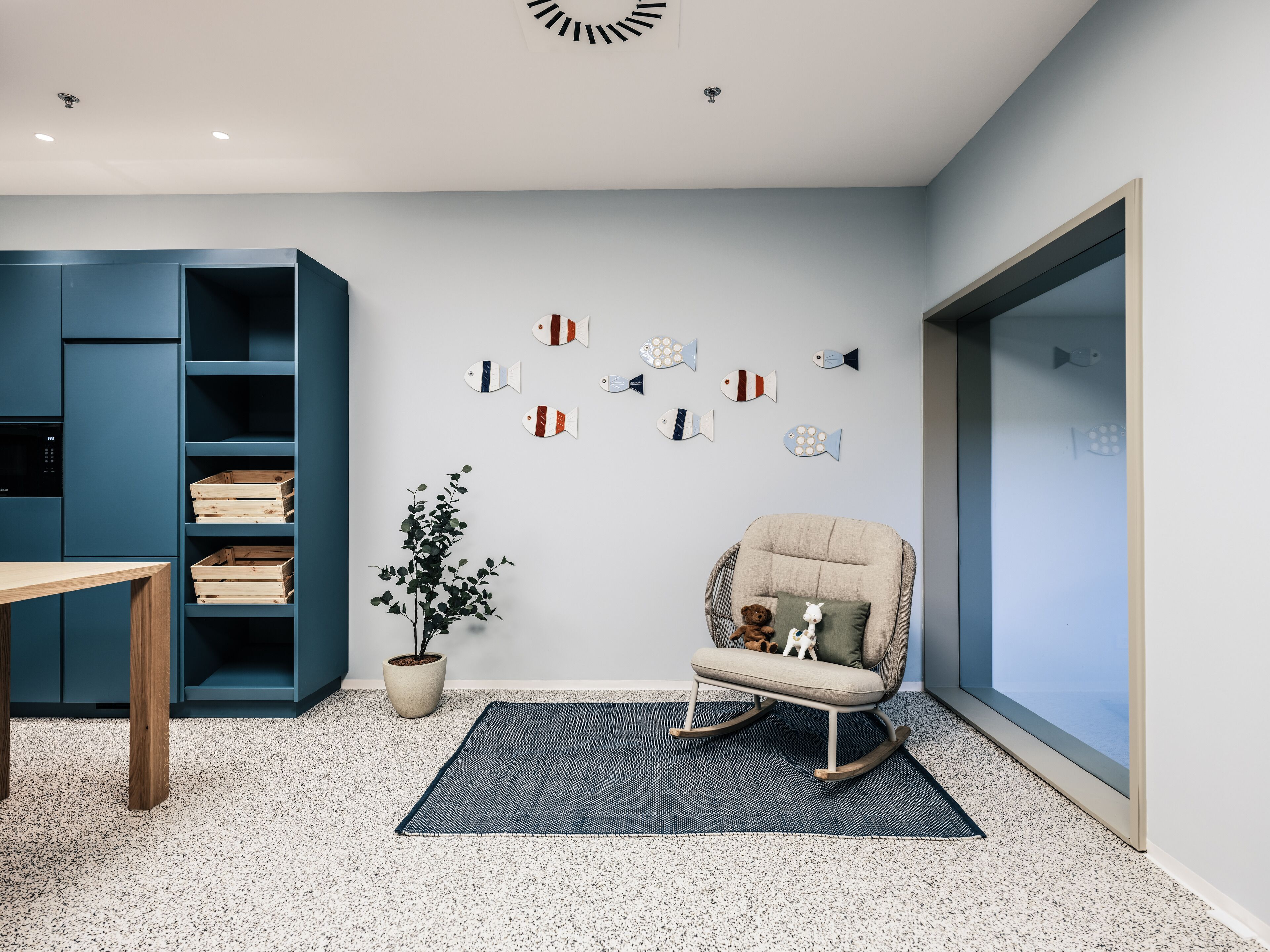 Un espacio de oficina moderno con un estante azul, una mecedora, una planta en maceta y adornos de peces en la pared.
