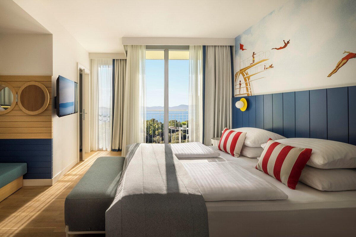 A hotel room with a large bed, a TV, a balcony with a view, and a wall mural.