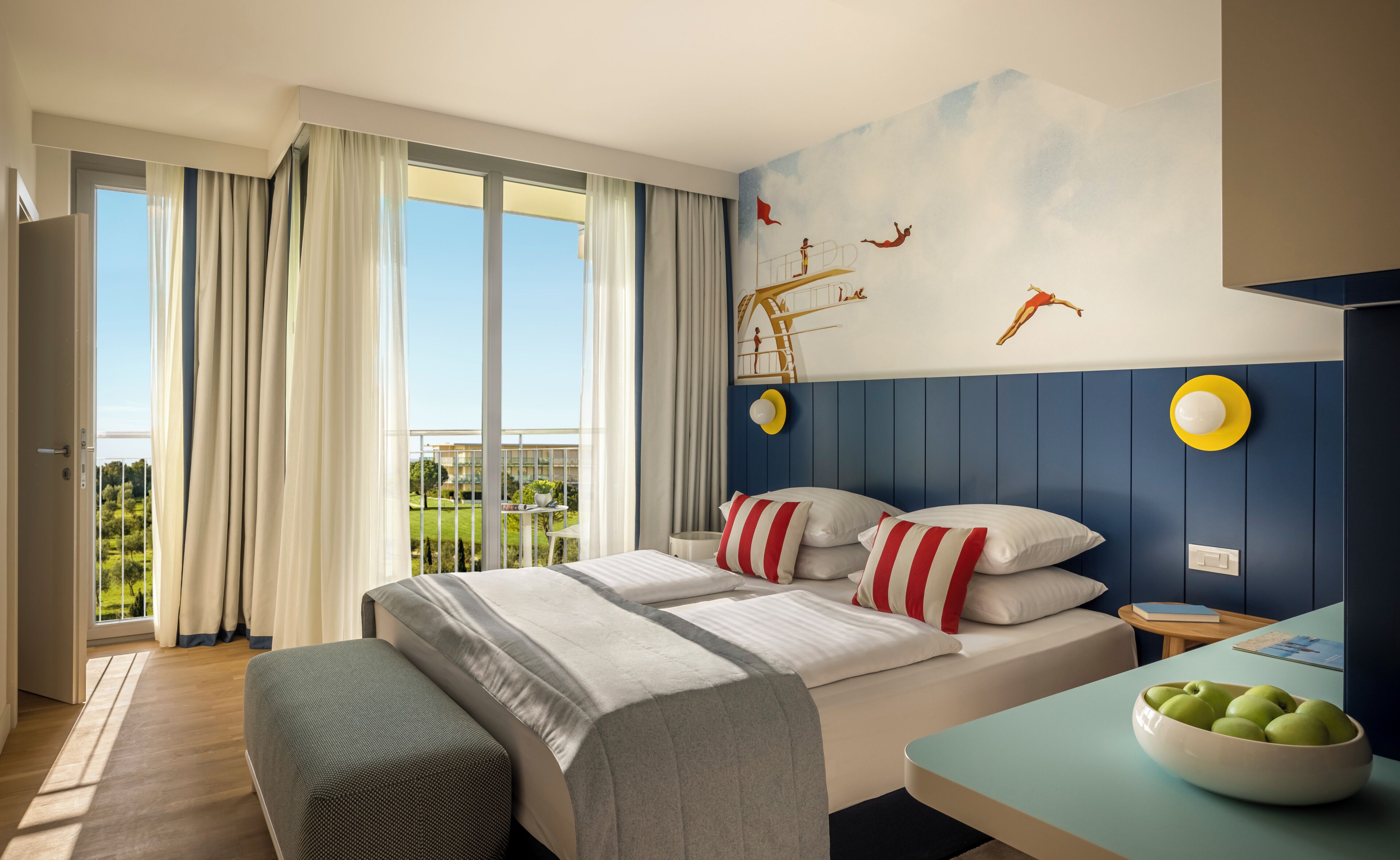 A hotel room with a bed, a desk with a bowl of apples, a view of greenery outside, and a wall mural.