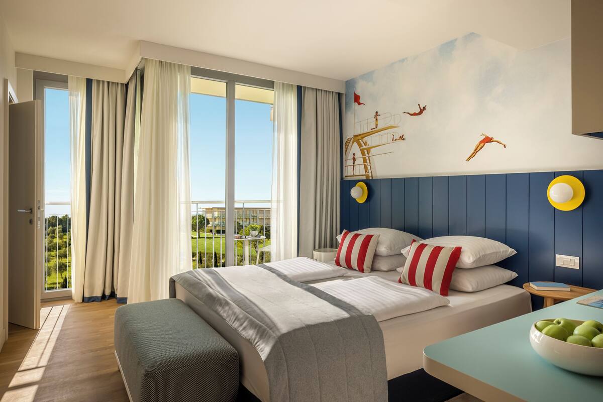 Ein Hotelzimmer mit Bett, Schreibtisch mit Apfelschale und Blick auf Grünfläche sowie einer Wandmalerei.