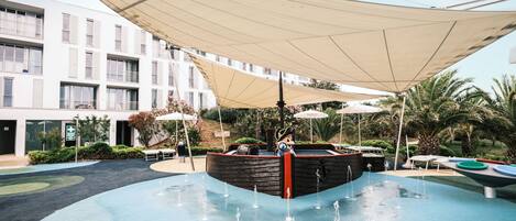 A swimming pool with a water fountain, surrounded by umbrellas and palm trees.
