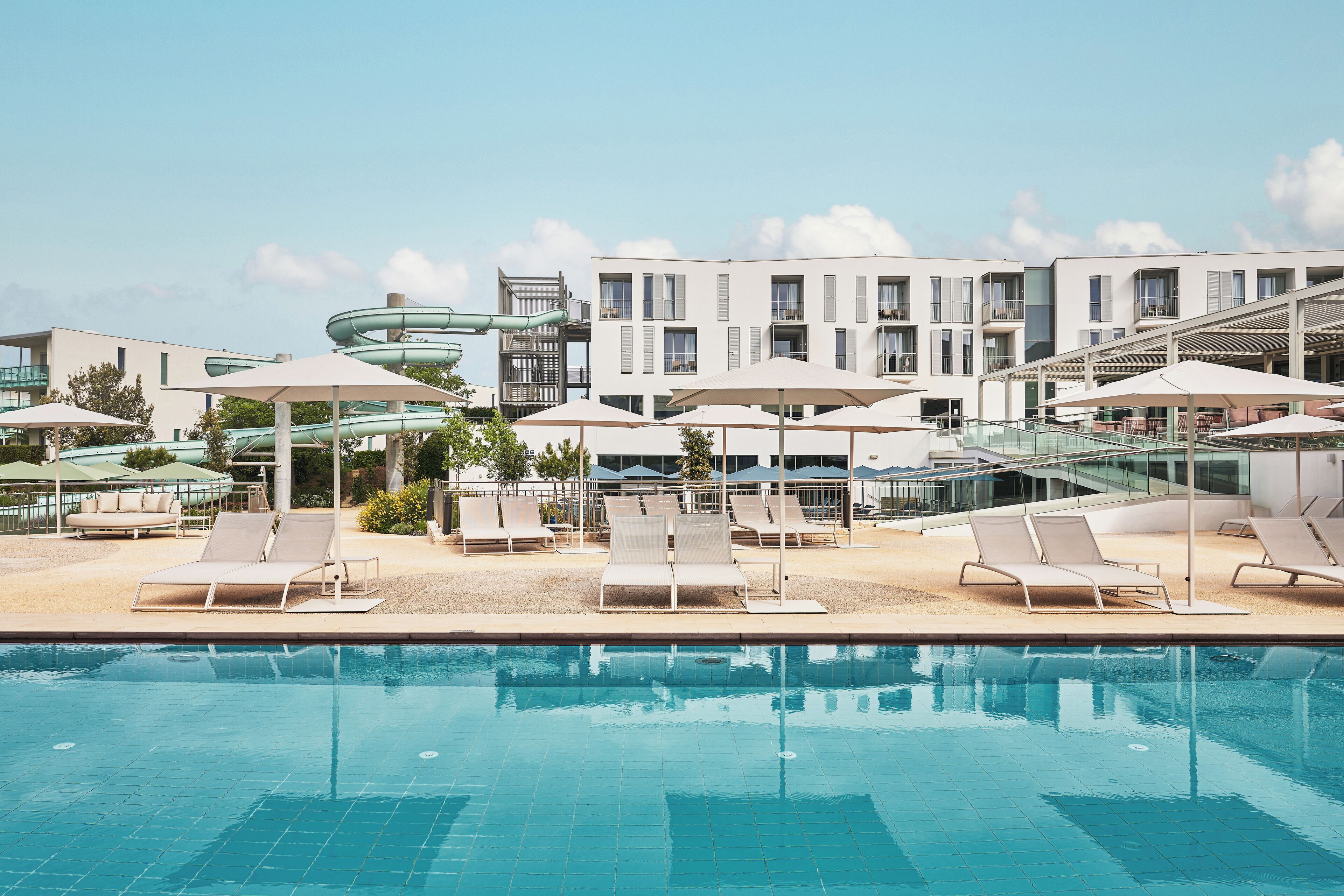 A modern hotel pool area with multiple lounge chairs and umbrellas, a water slide, and a multi-story building in the background.
