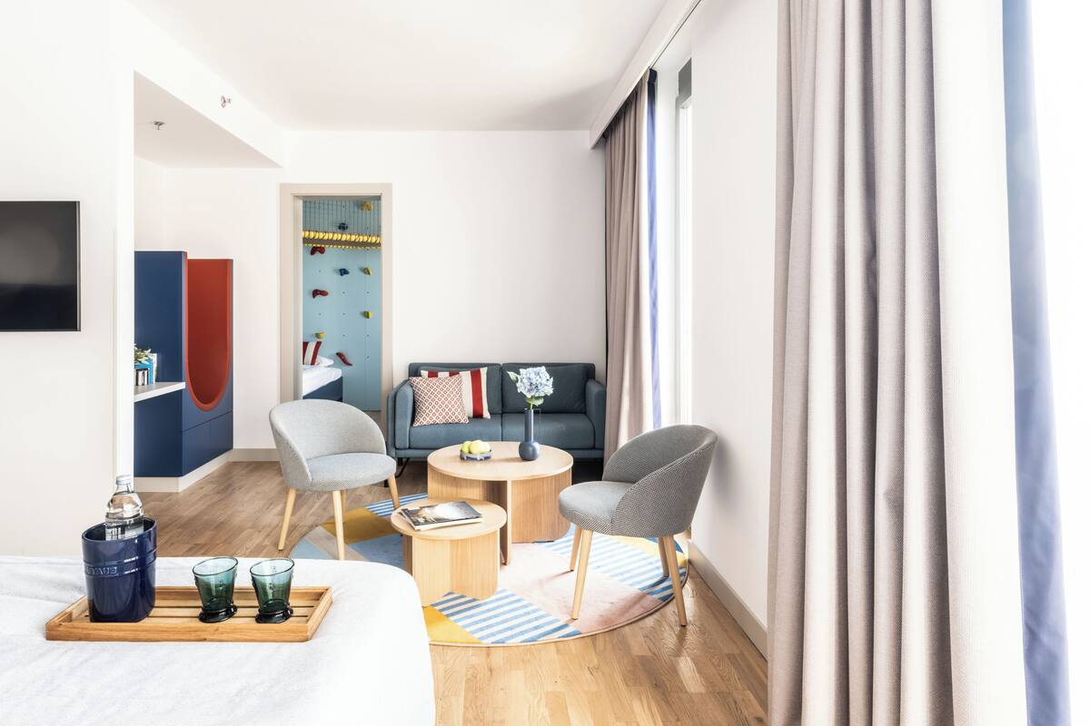 Ein Hotelzimmer mit einem Bett, zwei Sesseln, einem Sofa, einem kleinen Tisch, einem Fernseher und einem Fenster mit Vorhängen.