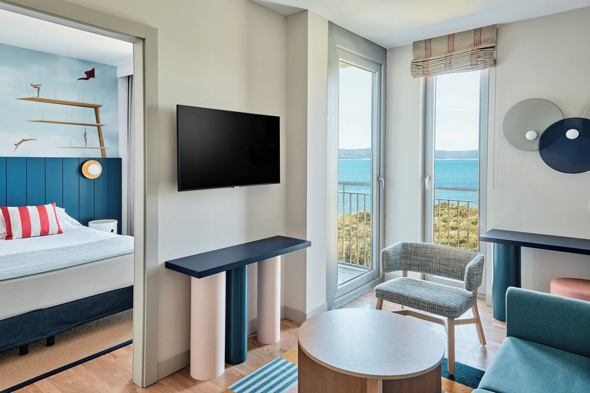 Ein modernes Hotelzimmer mit Bett, Fernseher, Schreibtisch, Sessel und einem Balkon mit Meerblick.