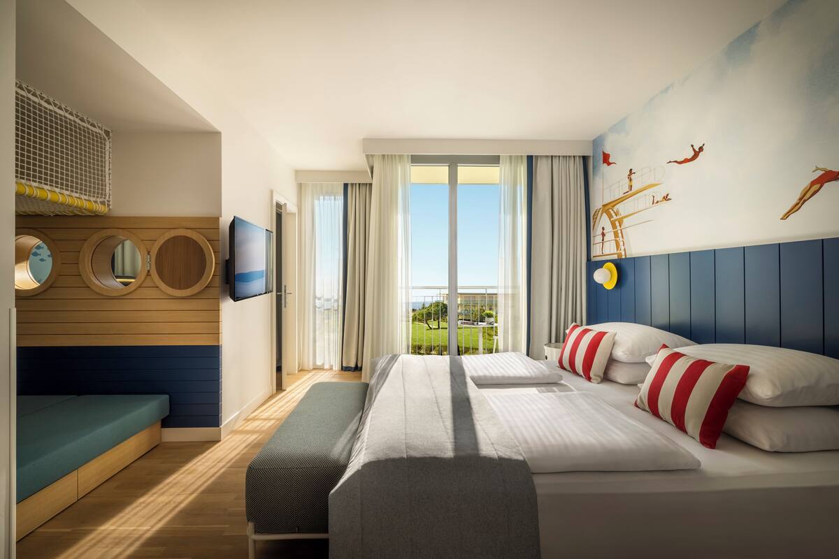 Ein Hotelzimmer mit einem Bett, zwei Nachttischen, einer Bank, einem Fernseher, einem Fenster mit Vorhängen und einem Wandbild.