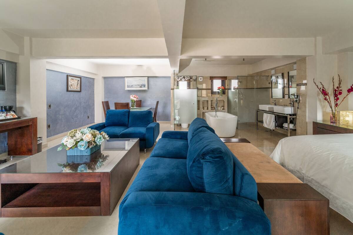 A modern hotel room with a large bed, a blue sofa, a wooden coffee table, and a bathroom with a bathtub and shower.