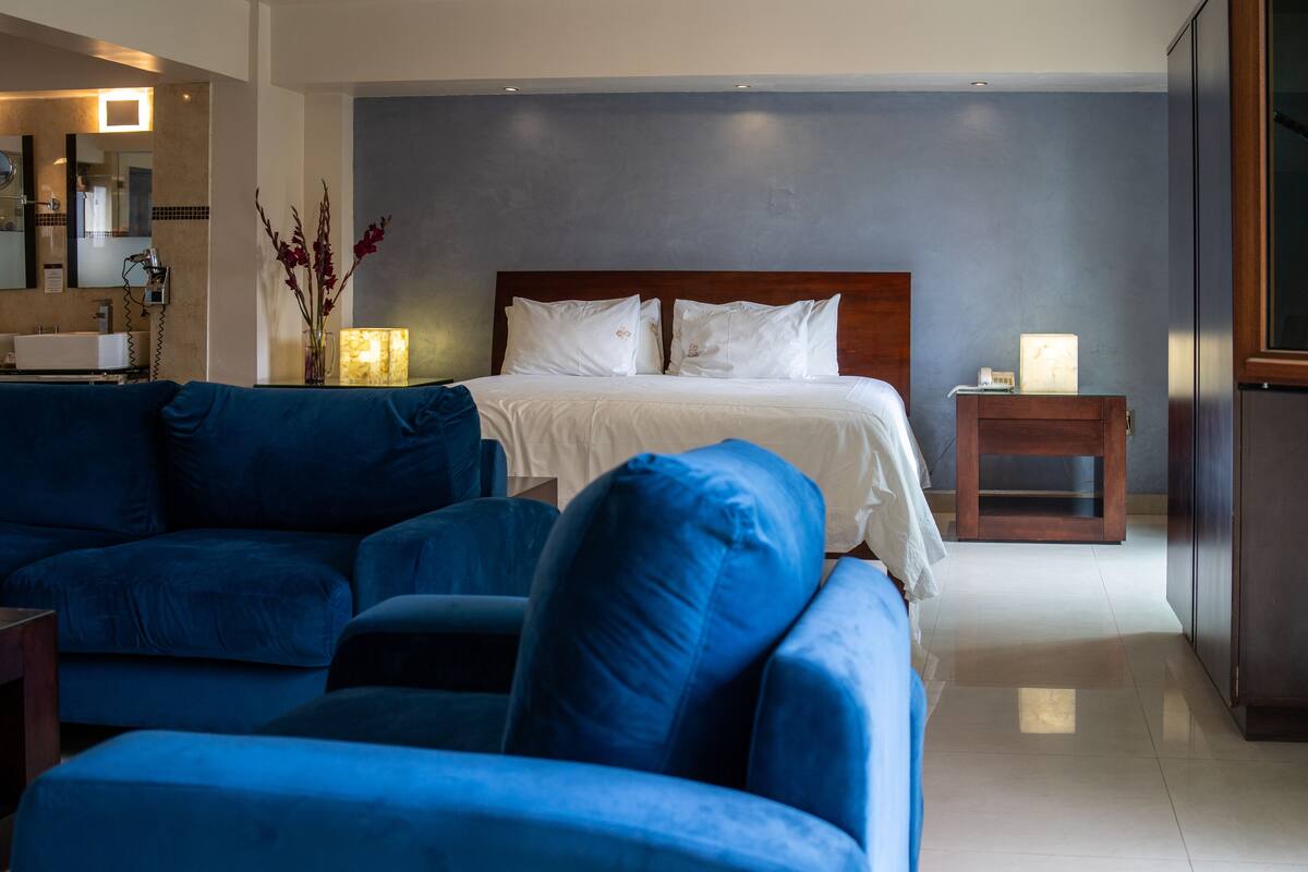 A modern hotel room with a large bed, a blue sofa, a bedside table with a lamp, and a mirror above the sink.
