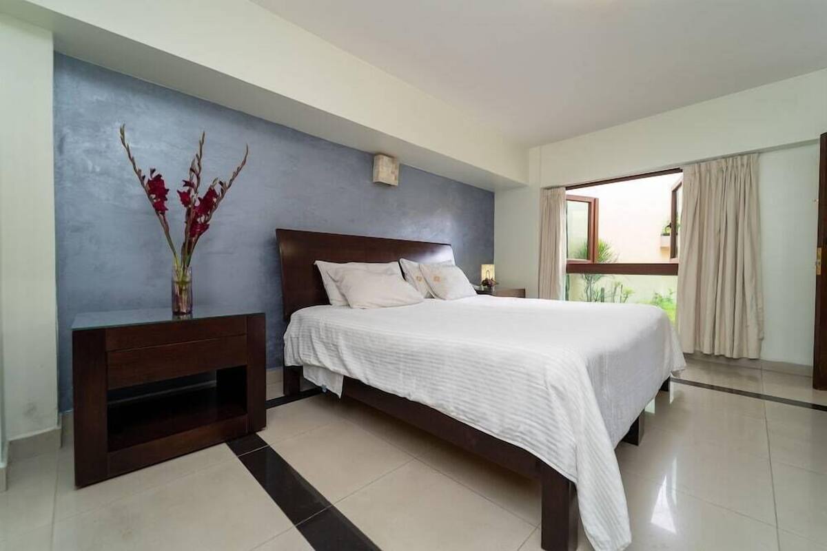 A bedroom with a bed, a nightstand, a vase with flowers, a window with curtains, and a view of greenery outside.