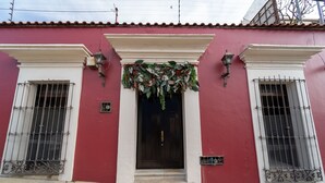 Un edificio rojo con una puerta blanca, dos ventanas con rejas y un adorno festivo sobre la puerta.