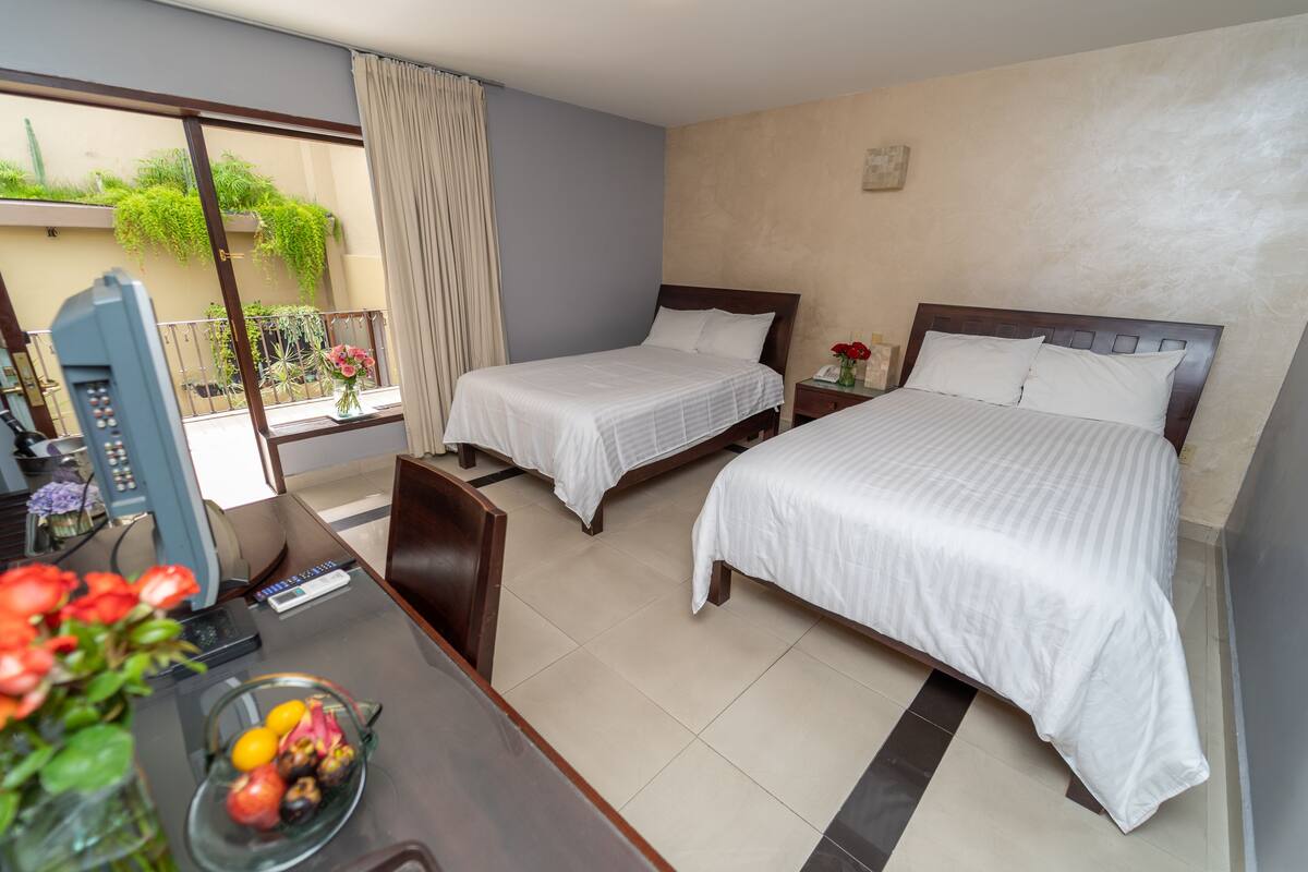 A hotel room with two single beds, a desk with a telephone, and a view of a balcony with plants.